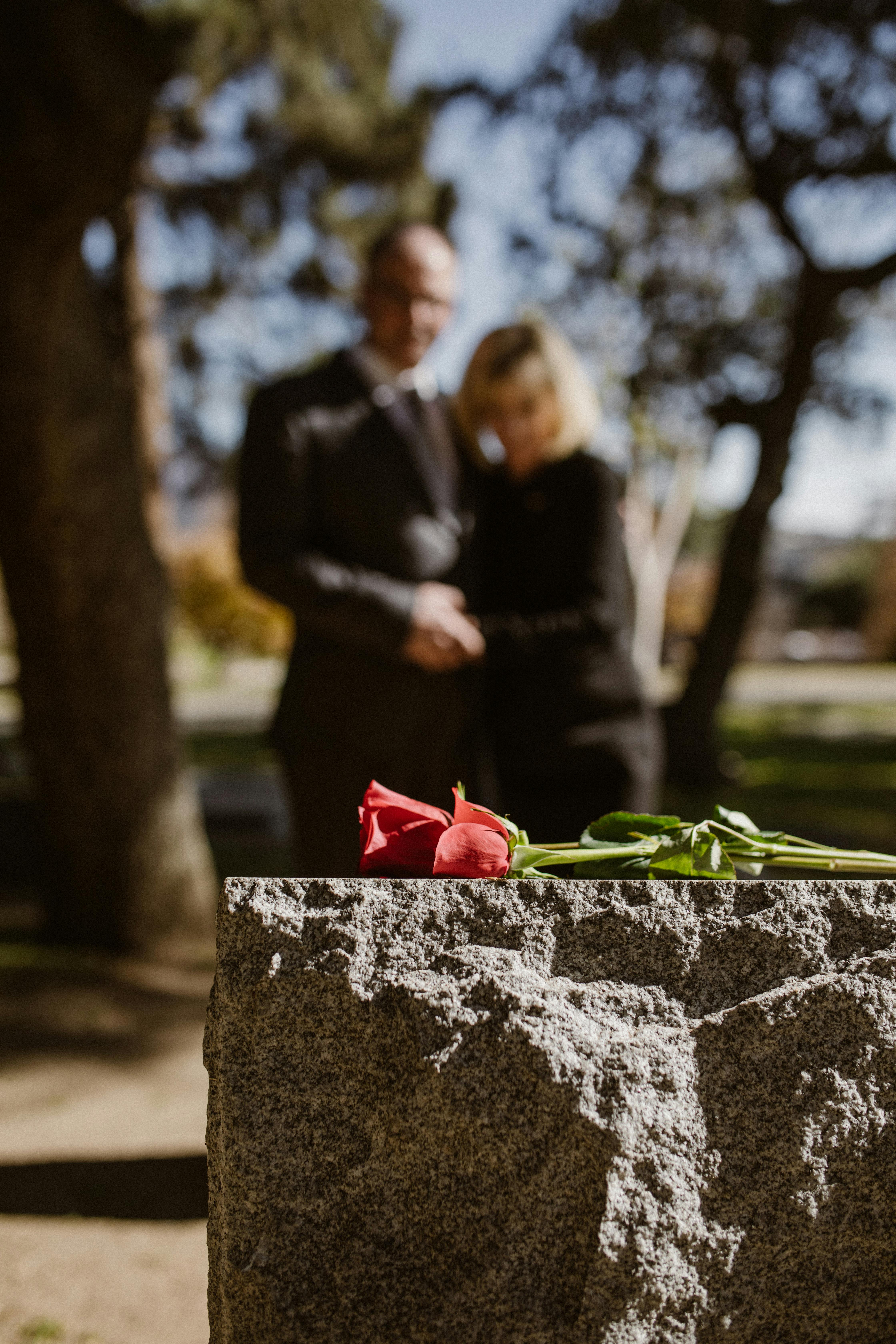 couple in black embracing next to headstone with red rose resting on top