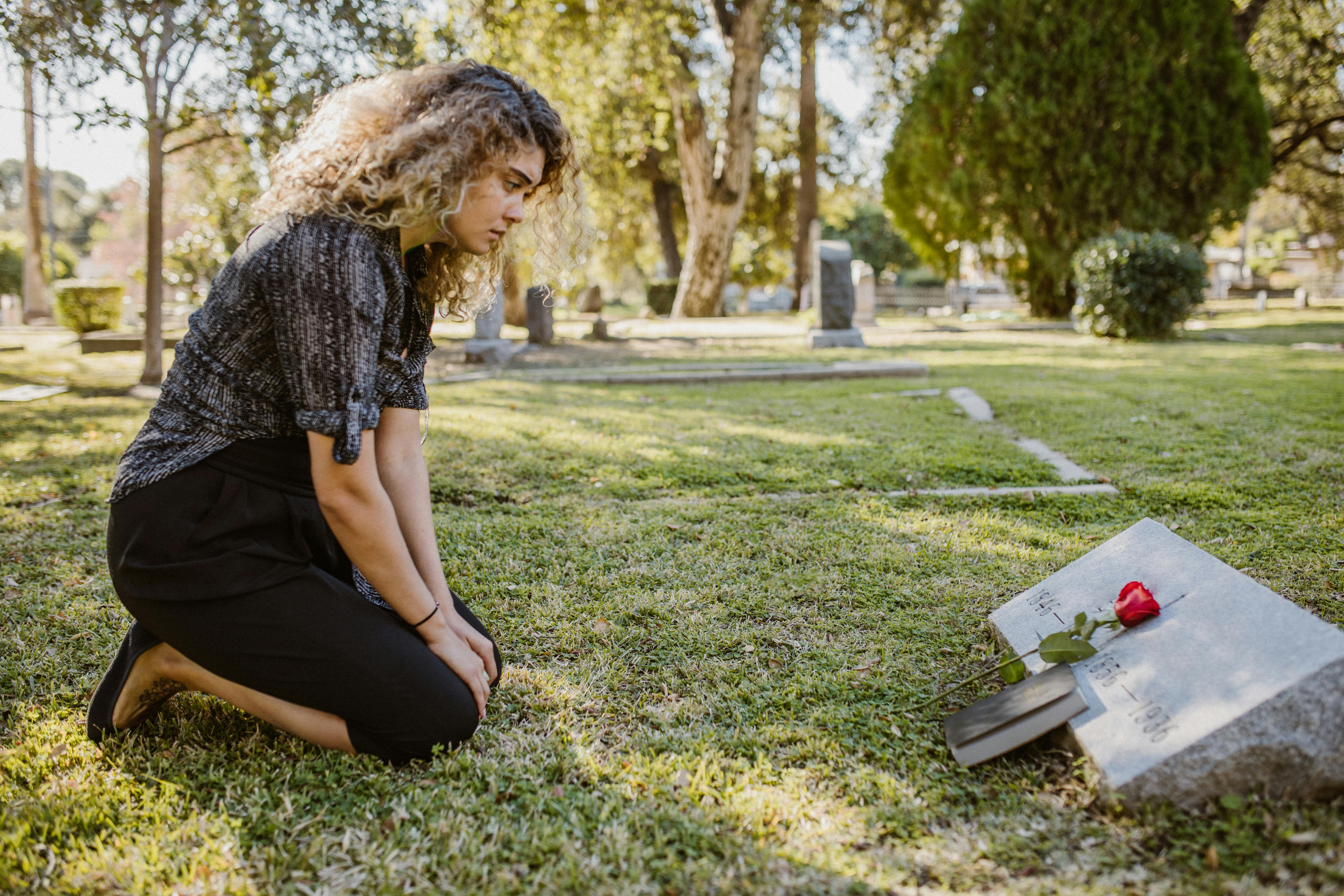 woman in black kneeling in front of a gravestone with Bible and red rose