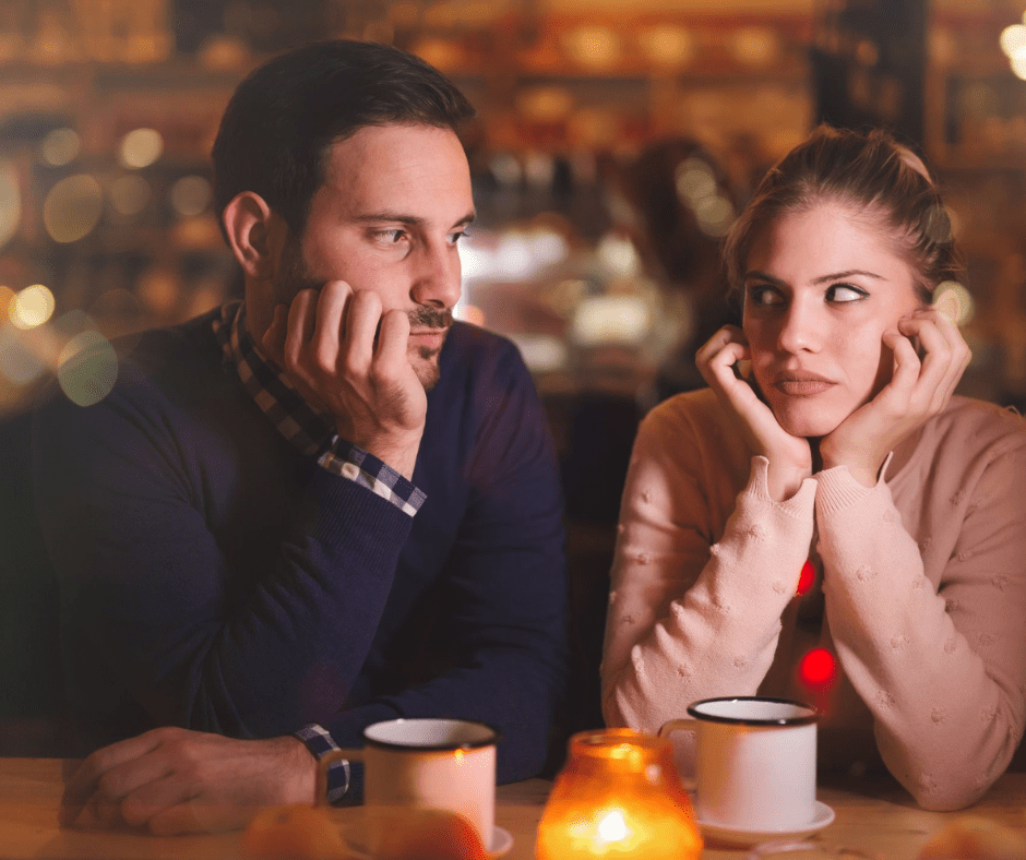 A man and woman sitting at a candlelit café table, both looking bored and frustrated, with cups of coffee in front of them.