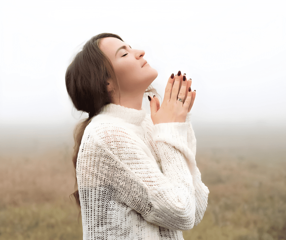 Woman in Nature Practicing Mindfulness and Gratitude Peaceful young woman with eyes closed and hands together in prayer, standing in a foggy field wearing a white sweater.