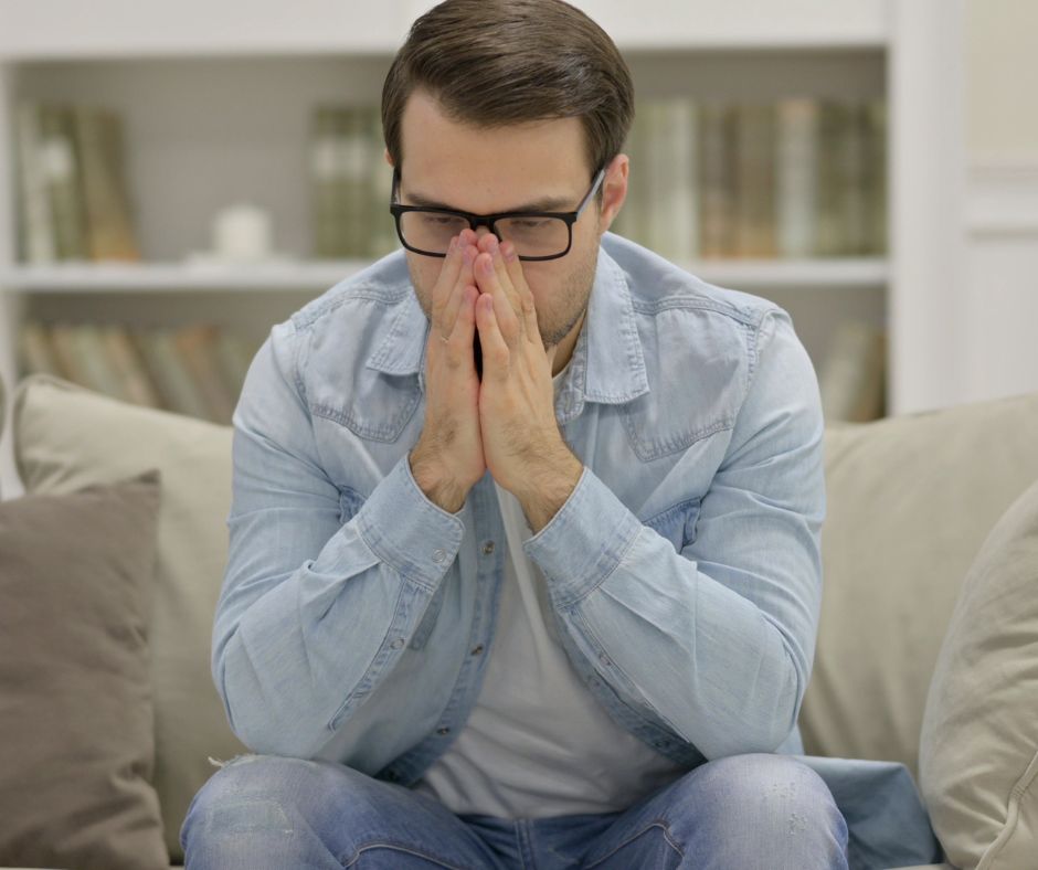 Young man covering her face with both hands, sitting by a window and expressing feelings of stress or overwhelm Young man covering her face with both hands, sitting by a window and expressing feelings of stress or overwhelm