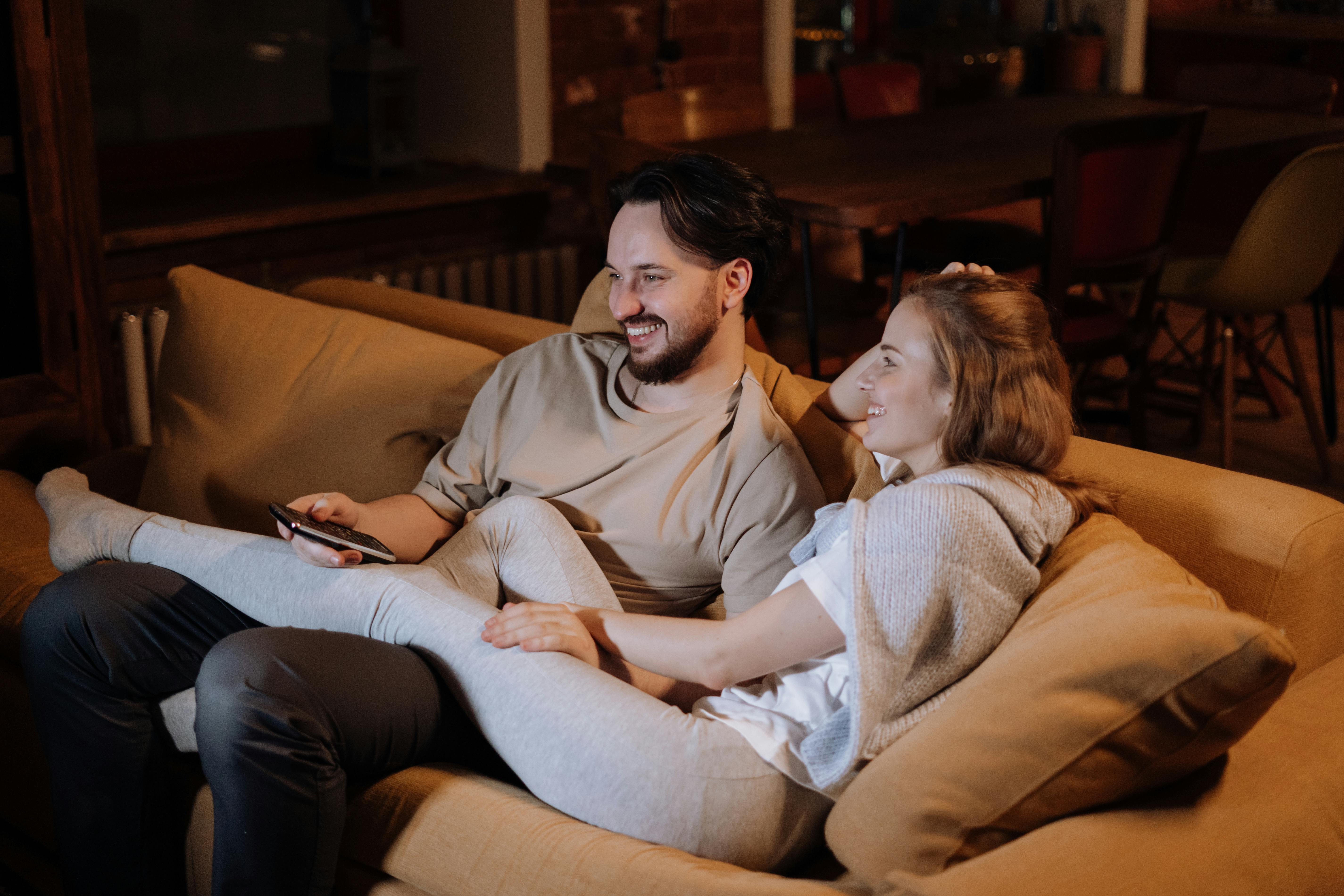 engaged-couple-relaxing-together-on-couch A smiling engaged couple relaxing together on a cozy couch at home, watching TV and enjoying quality time.